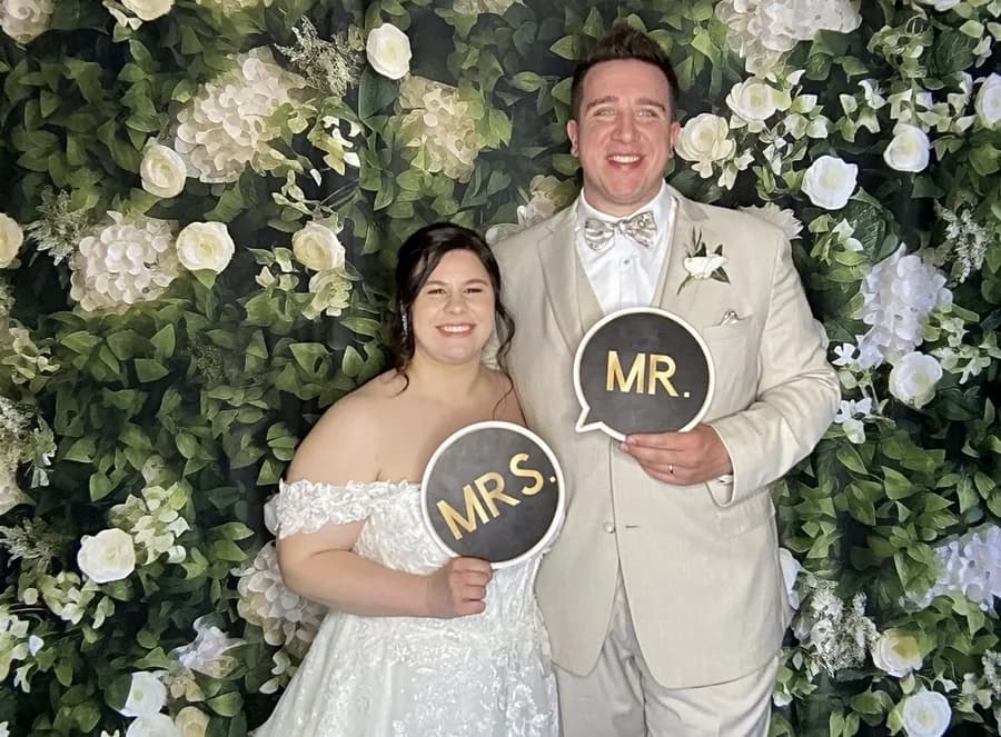 Anna and Robert posing with Mr and Mrs signs in the wedding photo booth against a floral backdrop